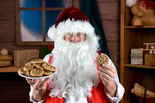Santa Claus in his workshop holding a plate full of fresh baked Chocolate Chip cookie