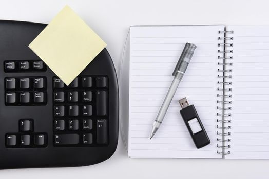Business Desk: Overhead view of a  notebook with a yellow post-it note, accompanied by a black computer keyboard with pen and thumb drive.