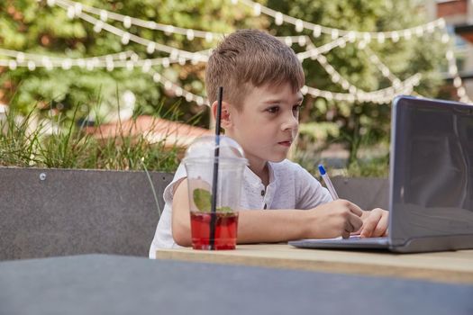 little schoolboy doing homework outdoors with laptop, writing notes in copybook