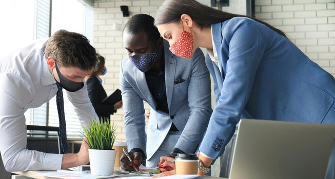 Business people wear preventive masks during epidemy in office. Collegues analysis finance growth success on meeting in office brainstorming, working on computers.