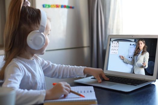 Smart small preschooler girl in headphones watch online lesson and communicate with teacher at home, little child in earphones study on Internet using laptop wireless connection.