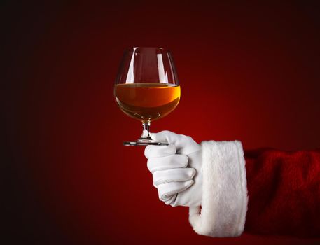 Closeup of Santa Claus holding a brandy snifter. Horizontal format on a light to dark red spot background.