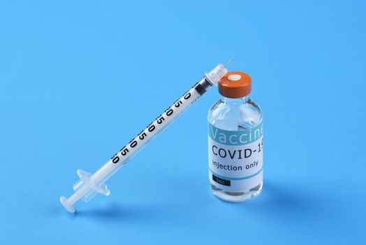 A vial of Covid-19 vaccine with a syringe leaning on the bottle on a blue background, with copy space.