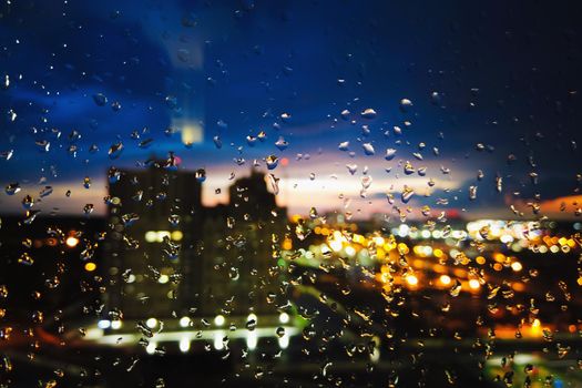 View through glass with raindrops on the night city. Sunset time, night city out of focus.