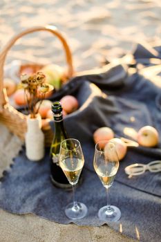 Focus on bottle of champagne and glasses, basket with fruits on plaid and beach in blurry background.