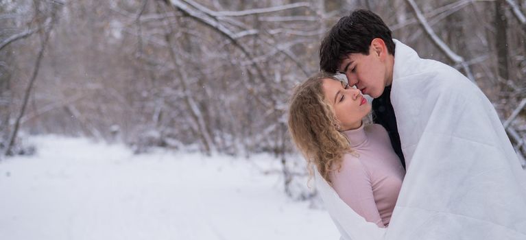 A young couple walks in the park in winter. The guy and the girl are kissing wrapped in a white blanket outdoors.