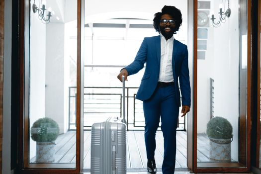 Black man in formal suit with packed suitcase entering hotel door
