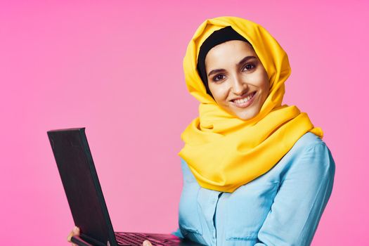 muslim woman in yellow hijab with laptop technology student pink background