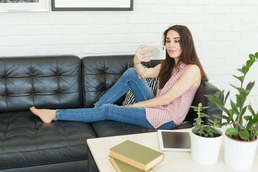 Technology, internet and people concept - young brunette blogger woman making photo using smartphone and lying on sofa at home