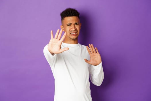 Image of african-american young man feeling uncomfortable, asking to stop, defending himself with raised arms and frowning, standing against purple background