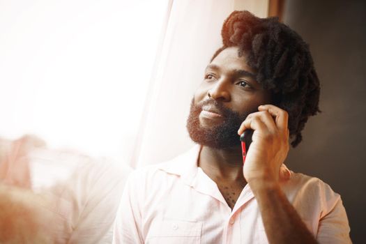 Close up photo of a black man in formal shirt talking on his phone near the window