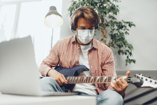 Young man plays guitar at home in medical mask. Coronavirus quarantine and self-isolation lifestyle concept