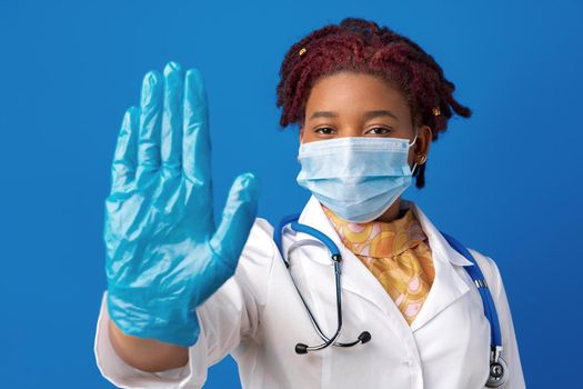 Portrait of african female doctor in lab coat with face mask and stethoscope against blue background