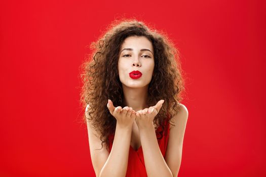 Woman rewards boyfriend with passionate wind kiss after romantic date. Portrait of sensual hot adult female in red elegant dress with curly hairstyle bending towards camera sending mwah flirty