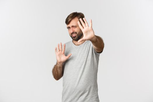 Portrait of displeased bearded man in gray t-shirt, defending himself from bright light, grimacing bothered, standing over white background