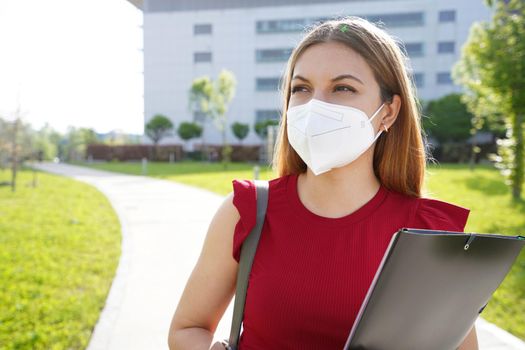 University student girl with FFP2 KN95 face mask holding folders looking to the side outdoors