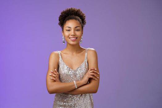 Confident wealthy attractive african woman in silver luxurious night dress hold hands crossed chest self-assured mighty pose smiling broadly enjoying rich life standing bossy blue background rejoice