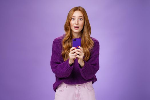 Impressed speechless cute redhead female actor receiving awesome surprising message on mobile phone open mouth from amazement and raising eyebrows astonished, posing over purple wall