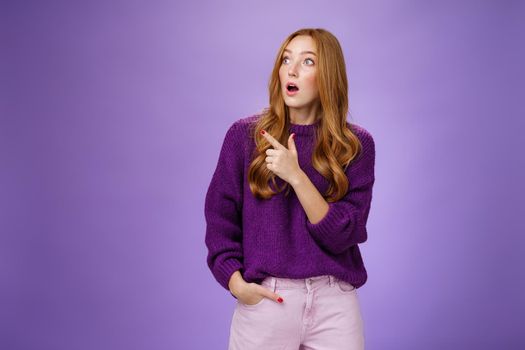 Girl surprised and thrilled as seing unbelievable scene. Portrait of impressed and shocked good-looking ginger woman in purple sweater gasping from amazement pointing, looking left intrigued