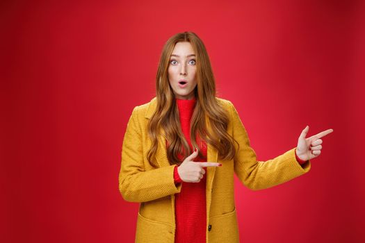 Speechless questioned and confused surprised redhead girl in yellow autumn coat pointing right open mouth curiously and staring at camera puzzled and clueless waiting explanation over red wall