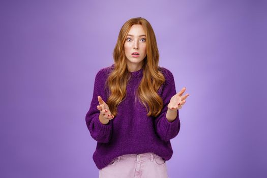 What wrong, I worried. Portrait of nervous and empathical young redhead woman feeling anxious raising hands questioned and looking at camera wondered expressing concern for friend over purple wall