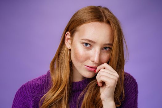 Ginger girl charmes with sensual romantic gaze. Portrait of confident flirting and attractive redhead female tilting head touching lips and looking daring at camera against purple background