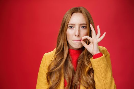 Girl putting seal on mouth making promise not tell anyone secret, sucking lips and holding finger near as zipping not spill words, looking serious and determined to keep surprise safe over red wall