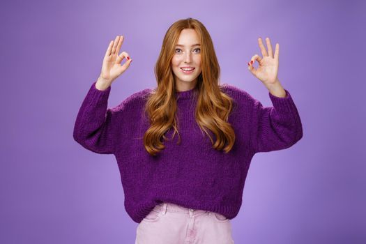 Fine I agree. Portrait of friendly and optimstic young 20s ginger girl in purple sweater raising hands with okay or ok gesture smiling in approval, liking cool product, giving recommendation