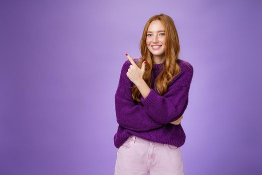 Charming friendly-looking bright redhead woman with freckles and makeup in purple warm sweater pointing at upper left corner and smiling delighted and cute at camera as showing cool place hang out