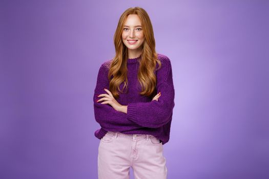 Portrait of pretty young and happy 20s ginger girl in purple sweater holding hands crossed over body smiling broadly, feeling self-assured as promoting project with confidence over violet wall