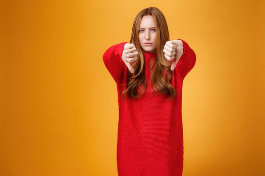 Oh no dislike. Portrait of upset and disappointed redhead female in red sweater frowning and pursing lips in dismay, showing thumbs down in disappointment and distress over orange background