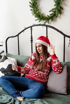 Young woman in red sweater and santa hat sitting on the bed using mobile phone texting or having video call