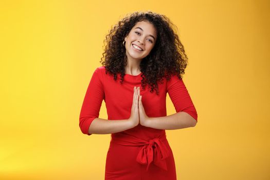 Welcome come inside. Portrait of friendly and polite good-looking female host in red dress with curly hair holding hands in namaste gesture tilting head and smiling as inviting guests in asian style