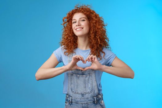 Sassy confident good-looking modern redhead curly woman raise head proud love tell girlfriend romantic heartwarming feelings present own heart smiling broadly standing blue background