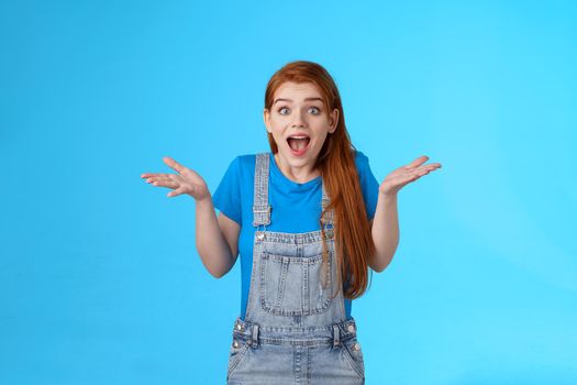 Excited girl describe awesome incredible news. Amazed friendly wondered redhead 20s woman show amazement and cheer, shrugging open mouth surprised, cannot believe what see, blue background
