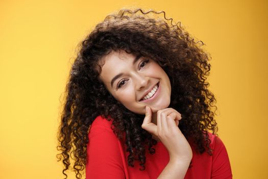 Close-up shot of charming flirty and silly curly-haired young woman making faces as trying get what wants, smiling tilting head on shoulder and touching lip standing cute over yellow background