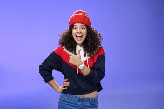 Portrait of enthusiastic and sociable european woman in warm hat and sweatshirt smiling delighted with amused grin as pointing at upper left corner impressed and astonished with awesome promotion