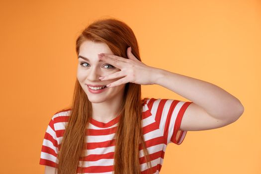 Charismatic lovely redhead lucky female, smiling cute, peek camera tender coquettish gaze, hold palm face, look through fingers, stand orange background optimistic, cheerful mood