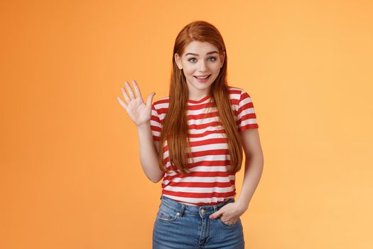 Hello how are you. Cute modest redhead newbie student female say hi, waving hand friendly, get know coworkers, smiling joyfully, introduce herself, welcome home, stand orange background