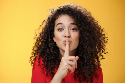 Shh please not tell secret. Cute and tender woman with curly hairstyle gently shushing with index finger on folded lips asking keep promise and remain silent, making surprise over yellow wall