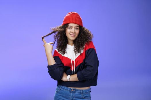 Stylish and snobbish arrogant curly-haired woman in warm beanie rolling curl on finger smirking and looking with contempt at camera, scorning person as being too cool over blue background