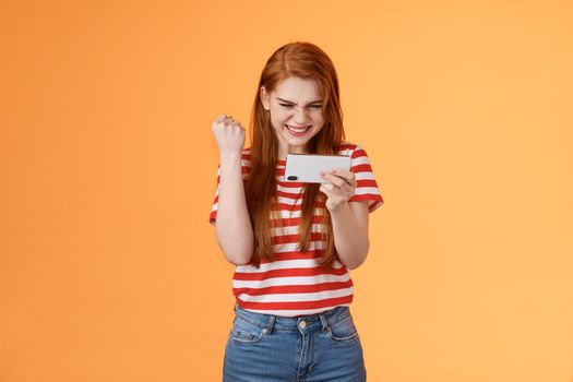 Pleased lucky redhead girl gamer redhead, fist pump satisfied, say yes excited stare display, hold smartphone horizontal, pass hard level playing awesome game use application, achieve victory