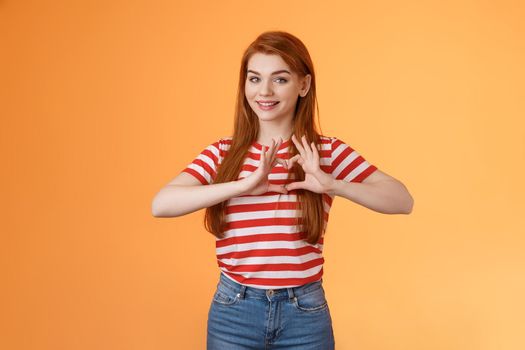 Cute lovely cheerful redhead girlfriend express love and cherish relationship, celebrating anniversary show heart sign, smiling tenderly, confess sympathy, stand orange background
