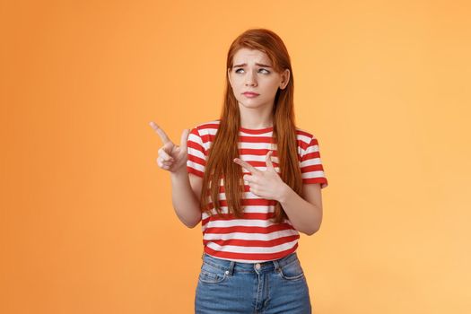 Complicated troubled cute young redhead girl puzzled facing tough difficult decision, look uneasy frowning perplexed hesitating, stare pointing upper left corner disappointed, orange background