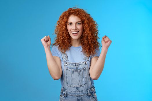 Hooray successful day. Cheerful chrismastic redhead girl summer dungarees clench fists joyfully smiling broadly determined achieve success, triumphing winning game standing upbeat blue background