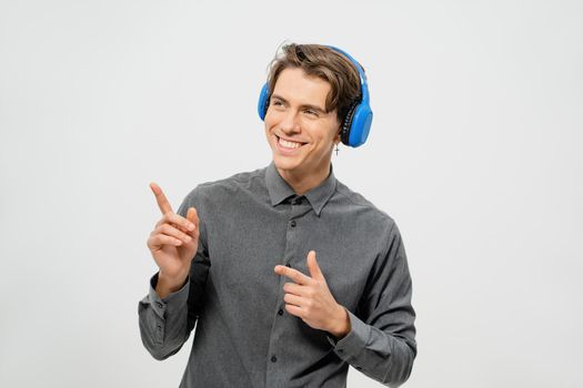 Happy smiling young guy in grey shirt standing listening music wearing blue wireless headphones pointing fingers sideways. Funny young guy listen to his favourite track or song