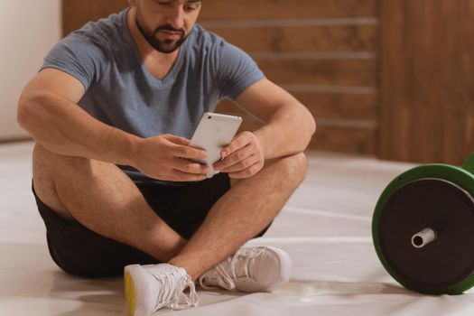 Body shot of handsome young man searching online or texting sitting on floor with smartphone, black and green tone fitness barbell, equipment for weight training concept. Healthy lifestyle concept