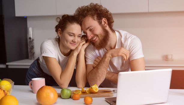 Beautiful young couple talking on video call using laptop. Young couple cooking healthy food in kitchen at home. Man leaned on girl smiling watching romantic movie