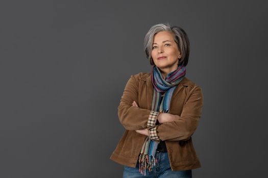 Pretty grey hair woman in casual isolated on grey background. Portrait of mature grey haired woman in a brown jacket looking positive on camera