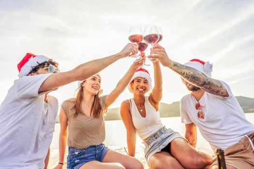 Group of multiracial happy young friends toasting with red wine glasses wearing Santa Claus red hat at sunset on a wooden pier in ocean sea resort for winter holidays vacations